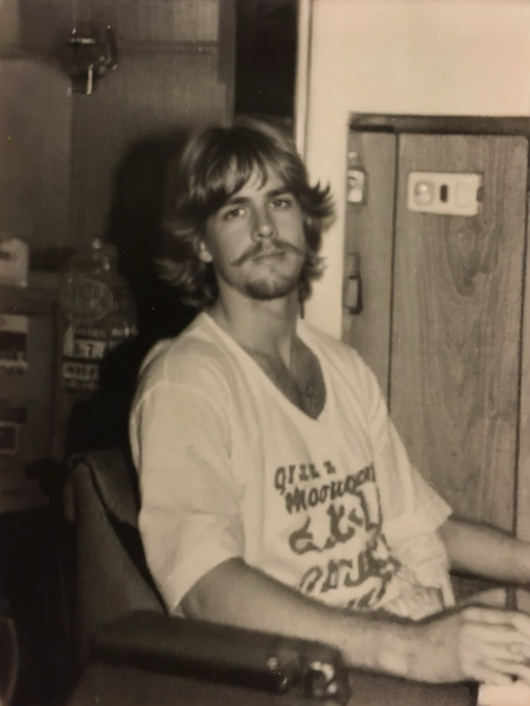 Black and white photo of a young man with 1970s-inspired long hair and mustache, wearing a vintage graphic t-shirt. A nostalgic throwback to retro fashion